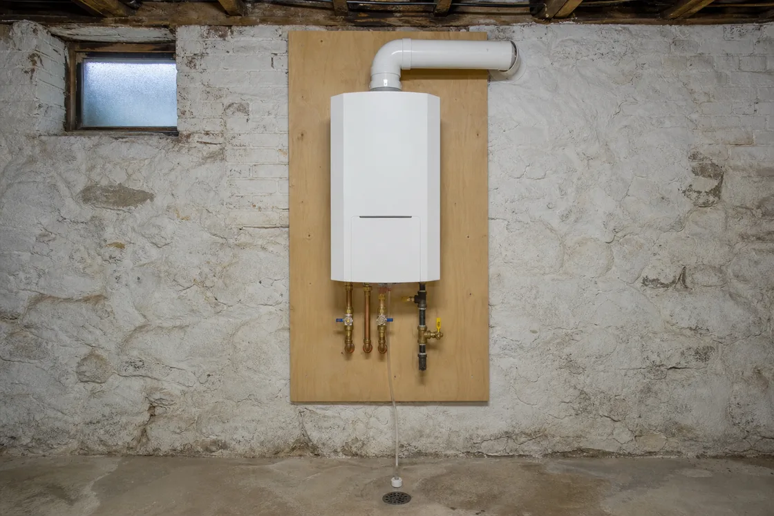 A wall-mounted tankless gas water heater on a backer board in a fieldstone basement, with isolation valves and a concentric vent
